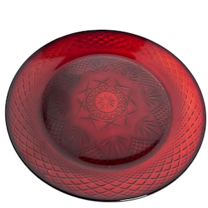 May include: A ruby red glass plate with intricate diamond and starburst patterns. The plate has a wide rim with a diamond pattern and a central design featuring a starburst pattern. The glass is transparent, allowing light to pass through.