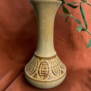 May include: A tall, slender ceramic vase with a speckled beige and tan finish. The base features a wide, rounded body decorated with a repeating embossed pattern of vertical, almond-shaped motifs, each containing a grid of horizontal lines.