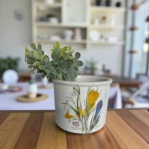 May include: A white ceramic planter with a floral design featuring yellow, purple, and white flowers, and green foliage. The planter is filled with green artificial plants and sits on a wooden table.