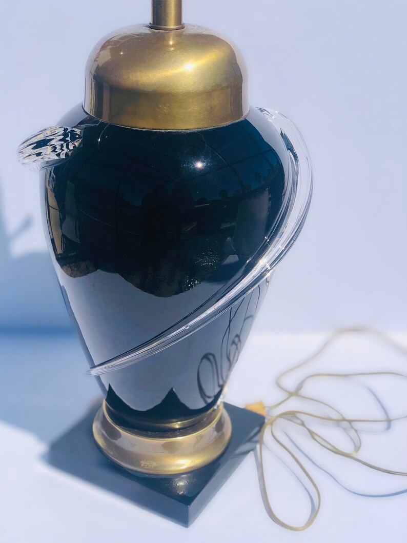 Puede incluir: Una l&aacute;mpara de mesa de vidrio negro con un elemento de dise&ntilde;o de vidrio transparente y una base de lat&oacute;n. La l&aacute;mpara tiene una base de m&aacute;rmol negro.