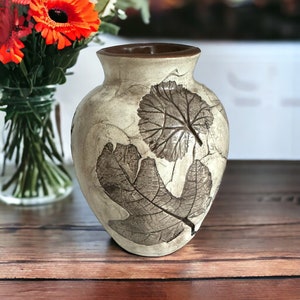 May include: A ceramic vase with a cream-colored base and a dark brown rim. The exterior features embossed dark brown leaf patterns with visible veins. The vase has a rounded body and a flared opening, showcasing a textured, matte finish.