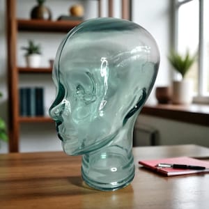 May include: A transparent, teal-colored glass head sculpture. The head is a stylized representation of a human face, with visible facial features. The glass has a smooth, reflective surface, and the sculpture is placed on a wooden surface.