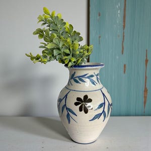 May include: A white ceramic vase with blue floral designs and a blue rim. The vase contains a green leafy plant. The vase has a brown flower in the center. The background is a light blue wooden panel.
