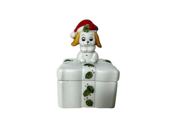 May include: White ceramic Christmas trinket box with a small dog wearing a Santa hat on top. The box is decorated with green holly leaves and red berries.