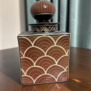 May include: A square ceramic canister with a brown and white arched scale pattern. The lid features a round, brown knob with white vertical lines and a decorative band of colorful, slanted geometric shapes in red, yellow, and blue.