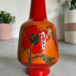 May include: A vibrant, orange ceramic vase with a red base and top. The vase is adorned with large, stylized red floral designs with green and black accents. The vase has a bulbous body and a slender neck, creating a striking decorative piece.