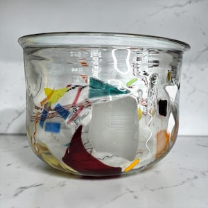 May include: A clear glass bowl with colorful abstract designs. The bowl features red, yellow, green, and blue shapes, with white and black accents. The bowl has a rounded shape and a wide opening, suitable for decorative use.