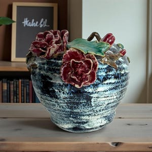 May include: A ceramic vase with a dark blue and white textured finish. The vase is adorned with red and green ceramic flowers. The vase is sitting on a wooden table.