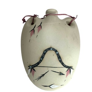 May include: A beige ceramic canteen with a leather strap and painted designs of feathers and an arrow. The canteen has a rustic, southwestern style.