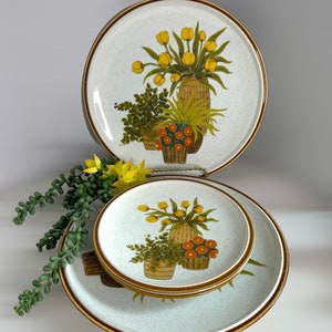 May include: A set of vintage ceramic plates with a floral design. The plates feature a speckled white background with a brown rim and a central illustration of yellow tulips, green foliage, and orange flowers in woven baskets.
