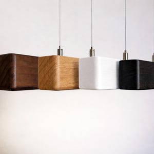 Peut inclure: Quatre lampes suspendues modernes avec des abat-jours carrés en bois ou peints, alignés. Les nuances sont marron, bois naturel, blanc et noir. Chaque lampe est suspendue par un fin fil d'argent.