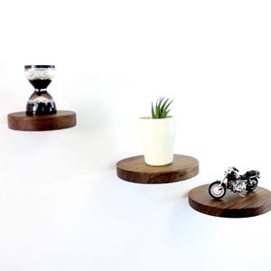Puede incluir: Tres estantes de madera montados en una pared blanca. El estante superior tiene un reloj de arena negro, el estante del medio tiene una pequeña suculenta en maceta y el estante inferior tiene una motocicleta de juguete negra.