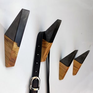 Puede incluir: Conjunto de cuatro perchas de pared geom&eacute;tricas de madera. Cada percha presenta una parte superior gris oscuro y una base de madera natural. Un cintur&oacute;n de cuero negro con hebilla plateada cuelga de una de las perchas.