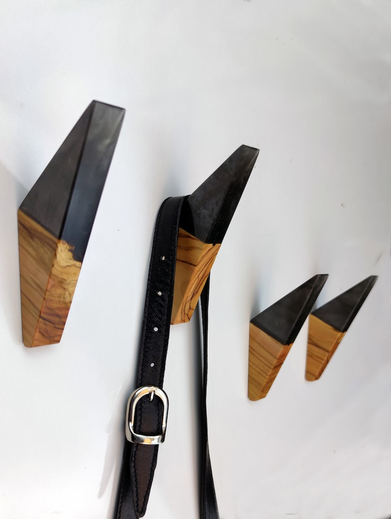 Puede incluir: Conjunto de cuatro perchas de pared geom&eacute;tricas de madera. Cada percha presenta una parte superior gris oscuro y una base de madera natural. Un cintur&oacute;n de cuero negro con hebilla plateada cuelga de una de las perchas.