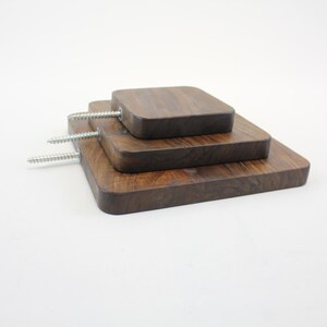Op de afbeelding: Drie houten wandplanken in verschillende maten, op elkaar gestapeld. De planken zijn gemaakt van donkerbruin hout en hebben een vierkante vorm. De planken zijn met schroeven aan de muur bevestigd.
