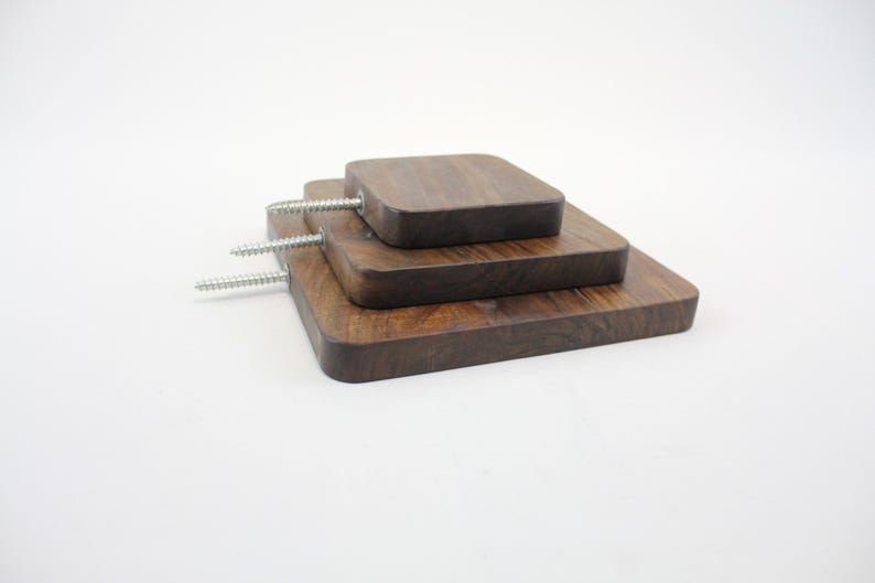 Op de afbeelding: Drie houten wandplanken in verschillende maten, op elkaar gestapeld. De planken zijn gemaakt van donkerbruin hout en hebben een vierkante vorm. De planken zijn met schroeven aan de muur bevestigd.