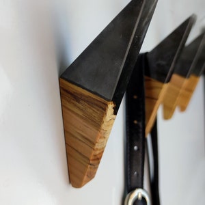 May include: Three triangular wooden wall hooks with black epoxy resin tops. The hooks are mounted on a white wall and a black leather belt is hanging on the leftmost hook.