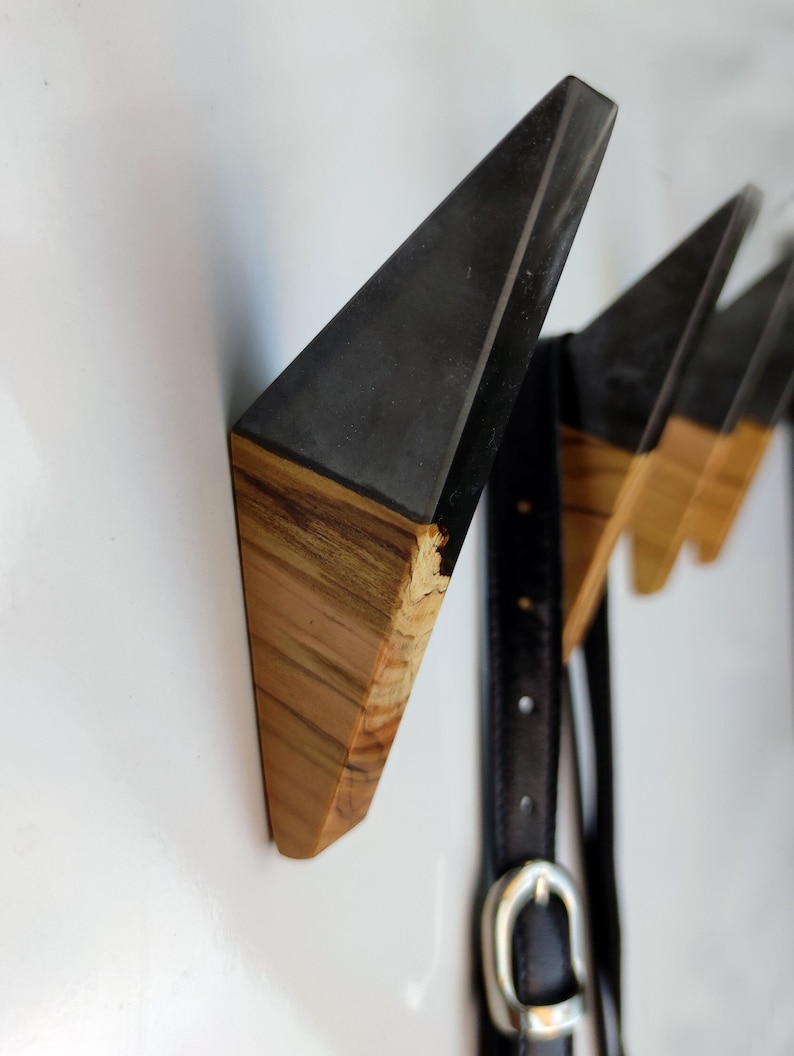 May include: Three triangular wooden wall hooks with black epoxy resin tops. The hooks are mounted on a white wall and a black leather belt is hanging on the leftmost hook.