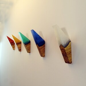 May include: Five wooden wall hooks with colorful resin inlays. The hooks are shaped like triangles and are arranged in a row from left to right. The colors of the resin are red, yellow, green, blue, and white.
