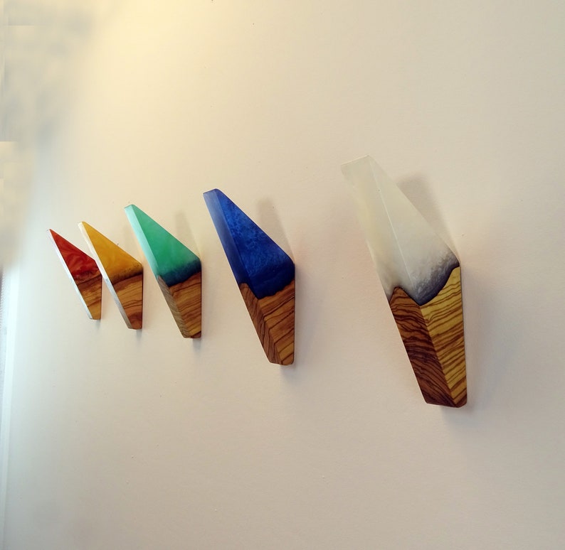 May include: Five wooden wall hooks with colorful resin inlays. The hooks are shaped like triangles and are arranged in a row from left to right. The colors of the resin are red, yellow, green, blue, and white.