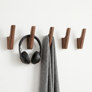 Könnte beinhalten: Fünf hölzerne Wandhaken mit geschwungenem Design, in einer Reihe angeordnet. Ein schwarzer Kopfhörer hängt an einem Haken, und ein grauer Schal an einem anderen. Die Haken sind an einer weißen Wand befestigt und bieten eine minimalistische Aufbewahrungslösung.