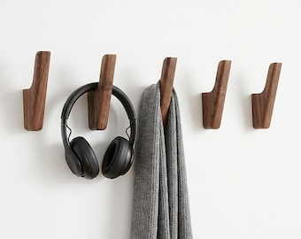 May include: Five wooden wall hooks in a row, each with a curved design. A pair of black headphones hangs from one hook, and a gray scarf from another. The hooks are mounted on a white wall, providing a minimalist storage solution.