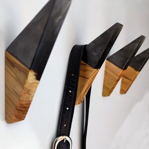Puede incluir: Juego de tres perchas de pared modernas con un dise&ntilde;o geom&eacute;trico. Cada gancho presenta una parte superior angular gris oscuro y una base de madera natural. Una correa de cuero negro con una hebilla plateada cuelga de un gancho.