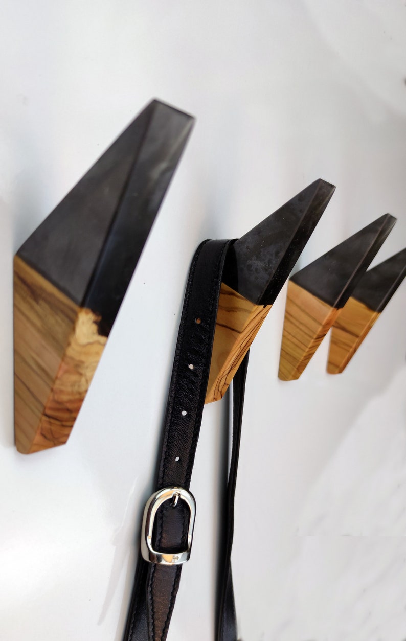Puede incluir: Juego de tres perchas de pared modernas con un dise&ntilde;o geom&eacute;trico. Cada gancho presenta una parte superior angular gris oscuro y una base de madera natural. Una correa de cuero negro con una hebilla plateada cuelga de un gancho.
