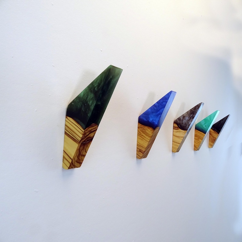 May include: Five wooden wall hooks with different colored resin inlays. The hooks are shaped like a triangle with a rounded point. The colors of the resin are green, blue, black, teal, and green.