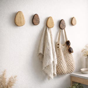 Puede incluir: Cinco perchas de pared de madera en tonos marrones variados están montadas en una pared blanca. Una toalla beige y un bolso tote tejido con gafas de sol cuelgan de dos de los ganchos. Los ganchos tienen forma orgánica.