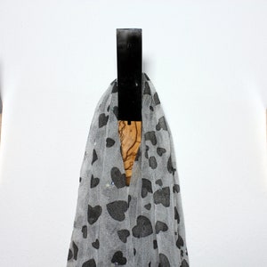 Puede incluir: Tres perchas de pared de madera con una parte superior de resina negra y una base de madera natural. Una bufanda gris transparente con patrones de corazones negros cuelga del gancho central. Las perchas est&aacute;n montadas en una pared blanca.