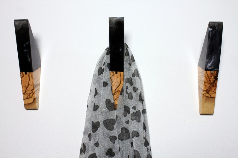 Puede incluir: Tres perchas de pared de madera con una parte superior de resina negra y una base de madera natural. Una bufanda gris transparente con patrones de corazones negros cuelga del gancho central. Las perchas est&aacute;n montadas en una pared blanca.