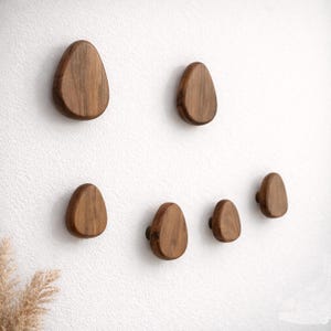 Puede incluir: Conjunto de seis perchas de pared de madera con forma de lágrima. Los ganchos son de madera de tonos cálidos y están montados en una pared blanca. Están dispuestos en un patrón atractivo, ofreciendo una solución de almacenamiento elegante.