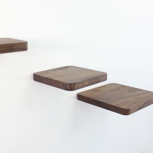 Op de afbeelding: Drie houten planken gemonteerd aan een witte muur. De planken zijn vierkant en gemaakt van donker hout. De planken zijn trapsgewijs geplaatst, waarbij de bovenste plank de kleinste is en de onderste plank de grootste.