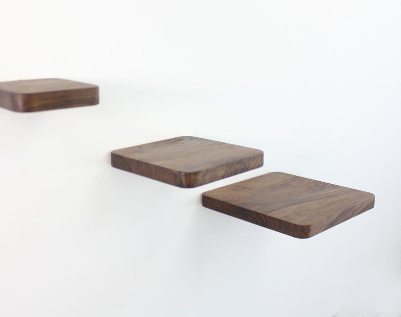 Op de afbeelding: Drie houten planken gemonteerd aan een witte muur. De planken zijn vierkant en gemaakt van donker hout. De planken zijn trapsgewijs geplaatst, waarbij de bovenste plank de kleinste is en de onderste plank de grootste.