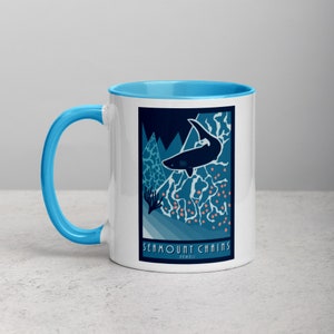 May include: White ceramic mug with a light blue rim and handle. The mug features a graphic of a shark swimming over a blue and pink seamount chain with the text "Seamount Chains, Hawaii".