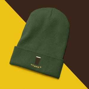 May include: A dark green beanie hat with a turned-up brim. The brim has an embroidered design of a brown glass with a white top and the words "VITAMIN G" in yellow.