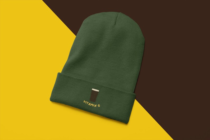May include: A dark green beanie hat with a turned-up brim. The brim has an embroidered design of a brown glass with a white top and the words "VITAMIN G" in yellow.