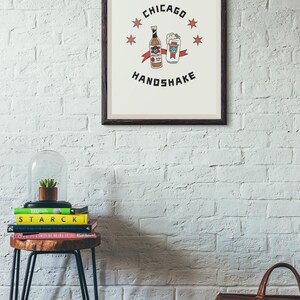May include: A framed print with a white background and a black border. The print features a cartoon illustration of a bottle of Mal&ouml;rt and a glass of beer with the text "Chicago Handshake" and five red stars.