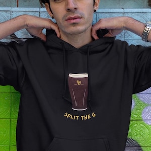 May include: Black hooded sweatshirt with a graphic of a pint of stout and the text "SPLIT THE G" in gold. The sweatshirt has a front pocket and a drawstring hood. The model is wearing the sweatshirt.