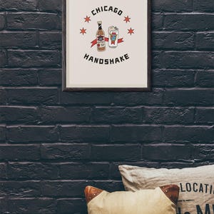 May include: A framed print with a white background and black text that reads "CHICAGO HANDSHAKE". The image features a cartoon drawing of a bottle of beer and a can of beer with red stars around the image.