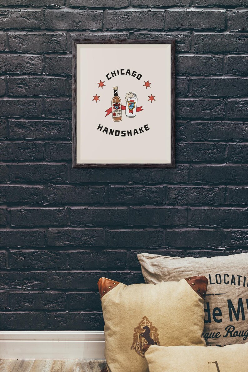 May include: A framed print with a white background and black text that reads "CHICAGO HANDSHAKE". The image features a cartoon drawing of a bottle of beer and a can of beer with red stars around the image.
