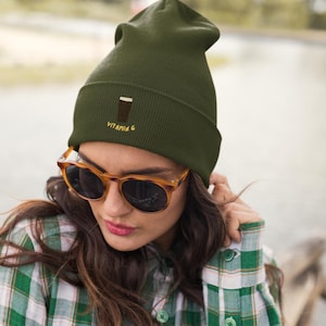 May include: An olive green beanie with the embroidered text "VITAMIN G" and a graphic of a drink. The person in the image is wearing tortoise shell sunglasses and a green and white plaid shirt.
