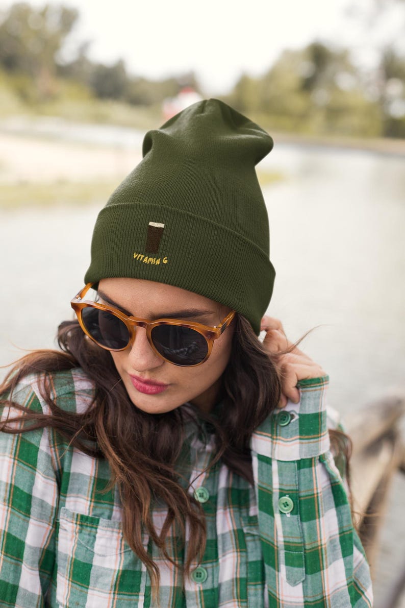 May include: An olive green beanie with the embroidered text "VITAMIN G" and a graphic of a drink. The person in the image is wearing tortoise shell sunglasses and a green and white plaid shirt.