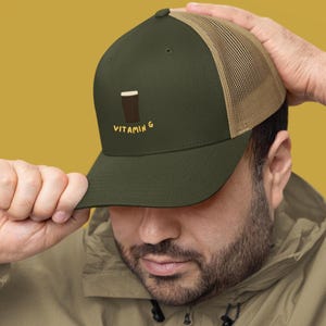 May include: Olive green trucker hat with a tan mesh back. The hat features a brown graphic of a beverage and the text "VITAMIN G" in yellow. The person in the image is wearing a tan jacket.