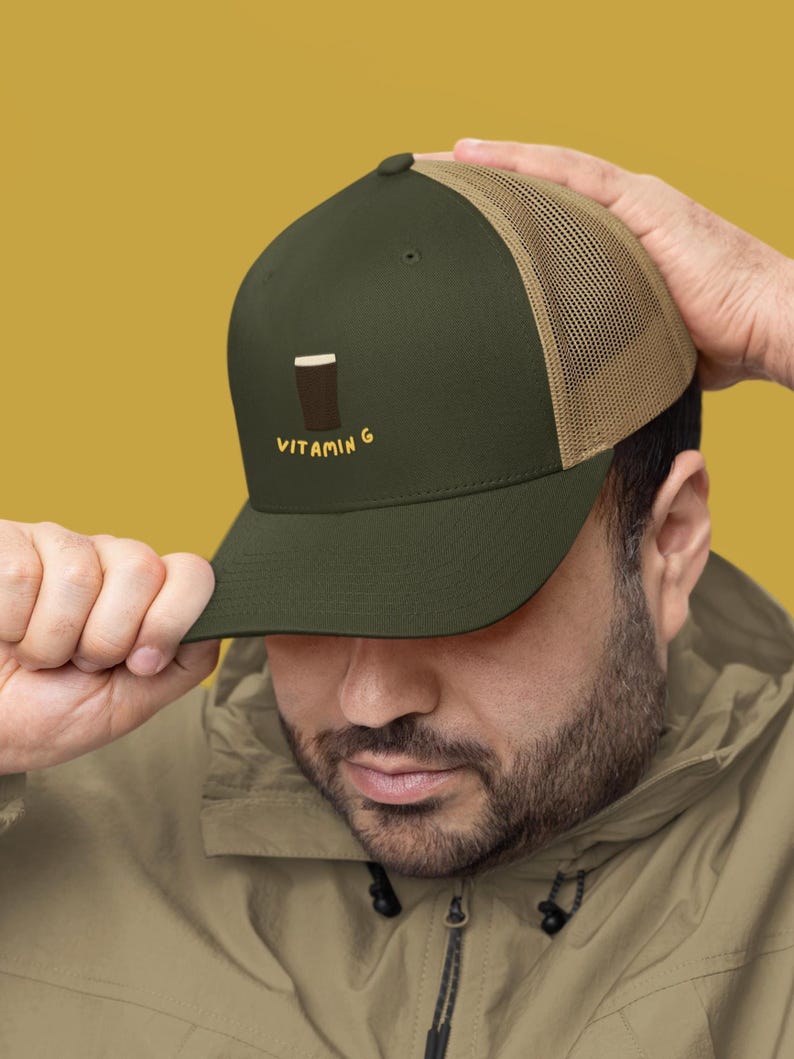 May include: Olive green trucker hat with a tan mesh back. The hat features a brown graphic of a beverage and the text "VITAMIN G" in yellow. The person in the image is wearing a tan jacket.