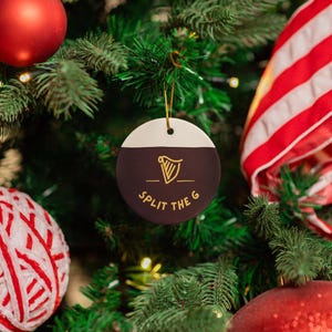 K&ouml;nnte beinhalten: Ein runder, schwarz-wei&szlig;er Weihnachtsbaumschmuck mit einem goldenen Harfendesign und dem Text "Split the G", der an einem gr&uuml;nen Weihnachtsbaum h&auml;ngt.