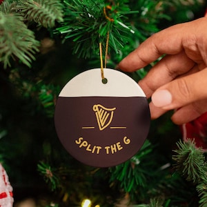 Op de afbeelding: Een ronde, witte en zwarte kerstbal met een gouden harpontwerp en de tekst "Split the G" in gouden letters.