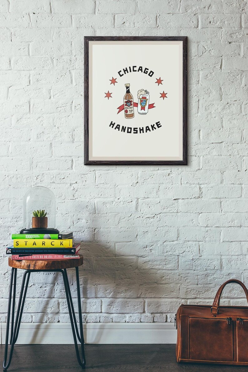 A framed Chicago Handshake art print hangs on a white brick wall above a small stool with books and a glass dome.