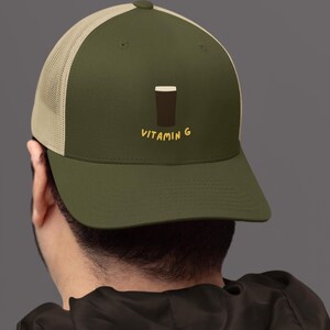 May include: Olive green trucker hat with a tan mesh back. The hat features a graphic of a dark brown pint of beer and the text "VITAMIN G" in yellow.